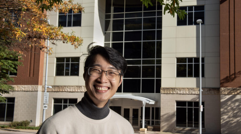 A smiling man in glasses stands in front of a large building.